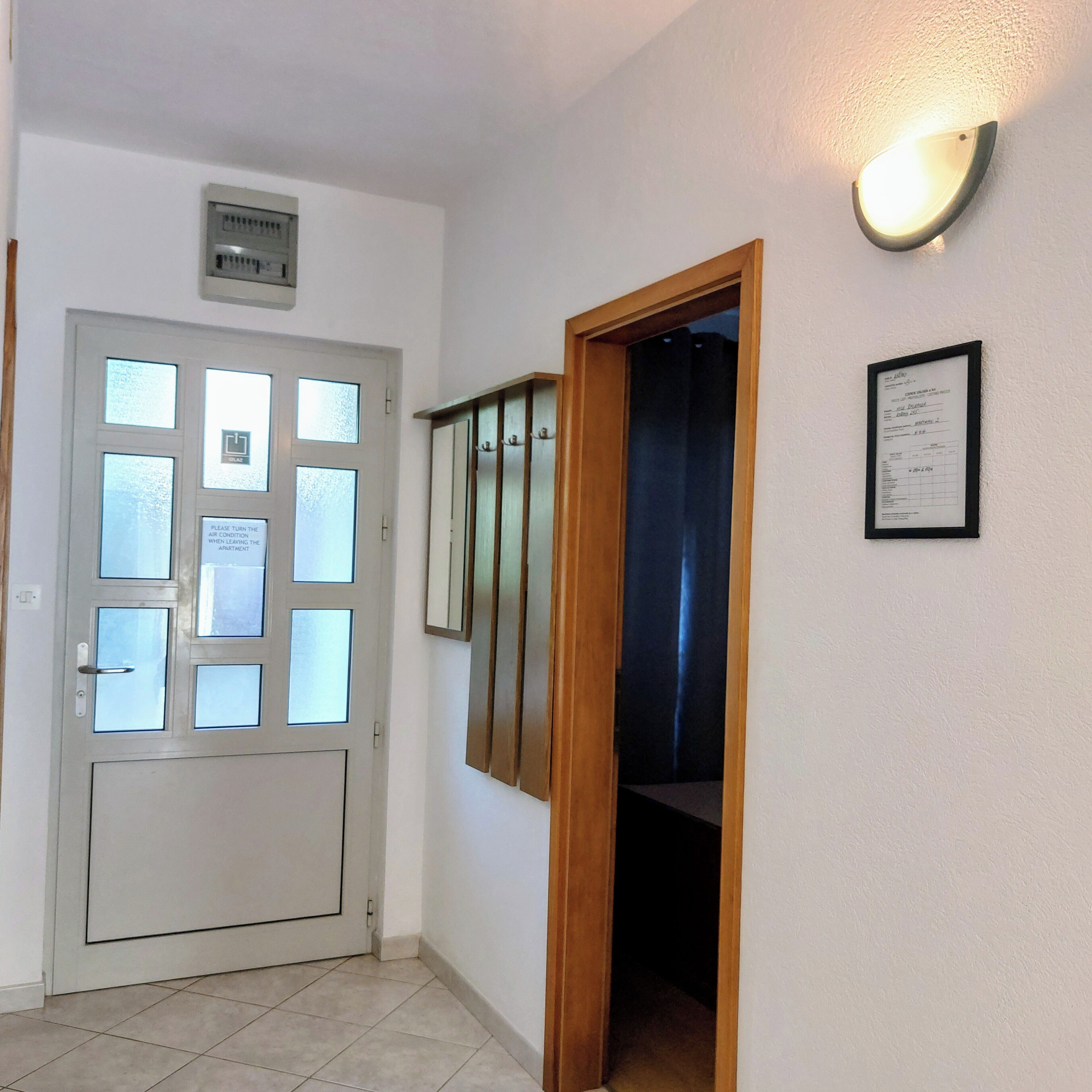 Ein Flur mit einer Tür, die mehrere Fenster aufweist, und einem Schild, das darauf hinweist, die Klimaanlage auszuschalten, wenn man das Zimmer verlässt. An der Wand hängt ein Aushang, und über der Tür befindet sich eine Leuchte.