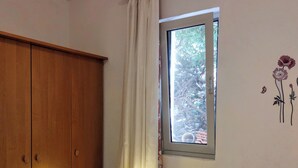 A bedroom with a wooden wardrobe, a bedside table with a lamp, a window with a view of trees, and a wall with a floral decal. - Apartment A2 dvije spavaće sobe, Garden Terrace 40 m2 with Outdoor Shower (Ražanj)