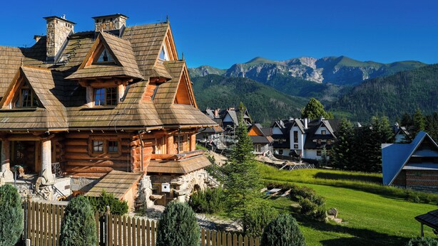 A traditional log cabin with a steeply pitched roof, surrounded by a wooden fence and lush greenery.