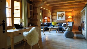 A cozy log cabin interior with a wooden desk, a comfortable sofa, a fluffy chair, and a fireplace.