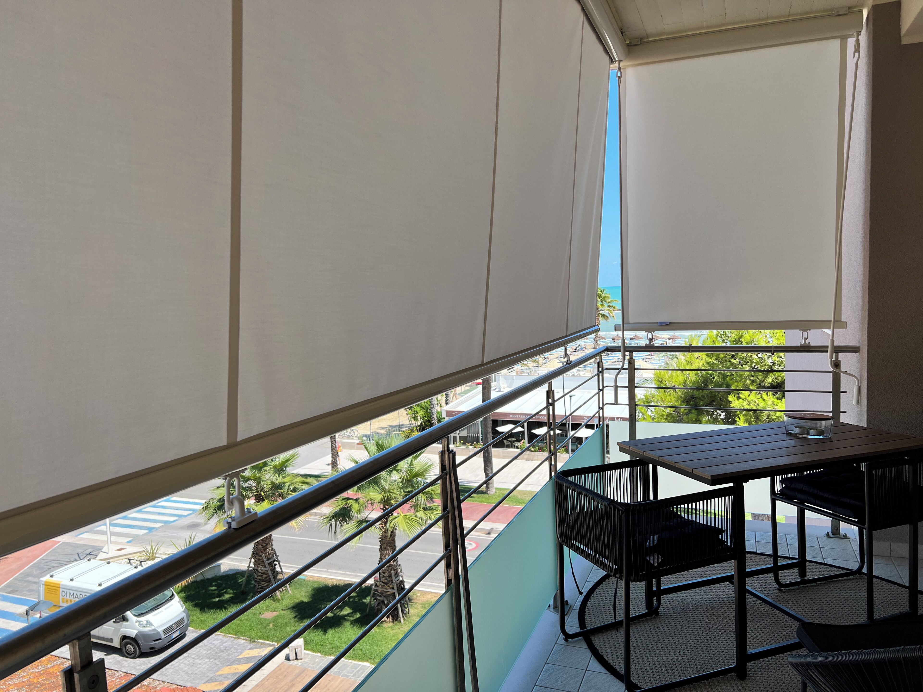 Un balcon avec une table et des chaises, une vue sur la rue et la verdure, et un fourgon blanc garé devant.