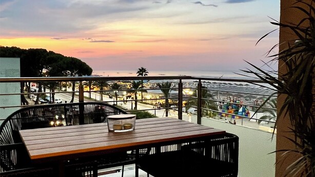 A balcony with a wooden table and chairs, overlooking a coastal area with a sunset view.