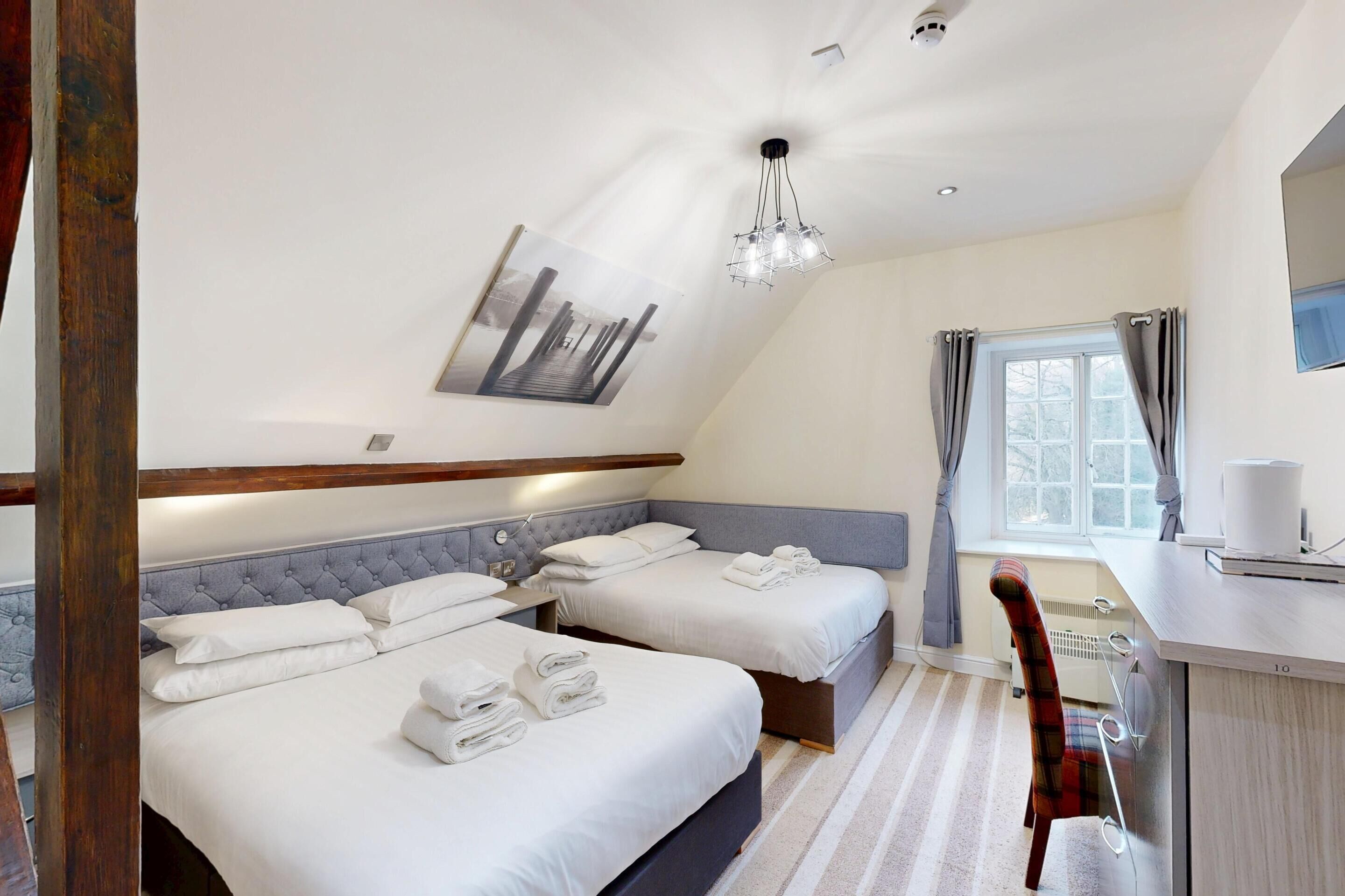 Habitación de hotel con dos camas, un escritorio, una silla, una ventana con cortinas y una lámpara en el techo.