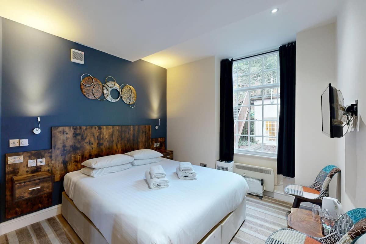 A modern hotel room with a large bed, a wooden headboard, a wall-mounted air conditioner, a window with curtains, and a small seating area with a chair and table.