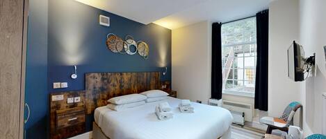 Habitación de hotel moderna con una cama grande, cabecera de madera, aire acondicionado empotrado, ventana con cortinas y un pequeño rincón de descanso con una silla y una mesa.