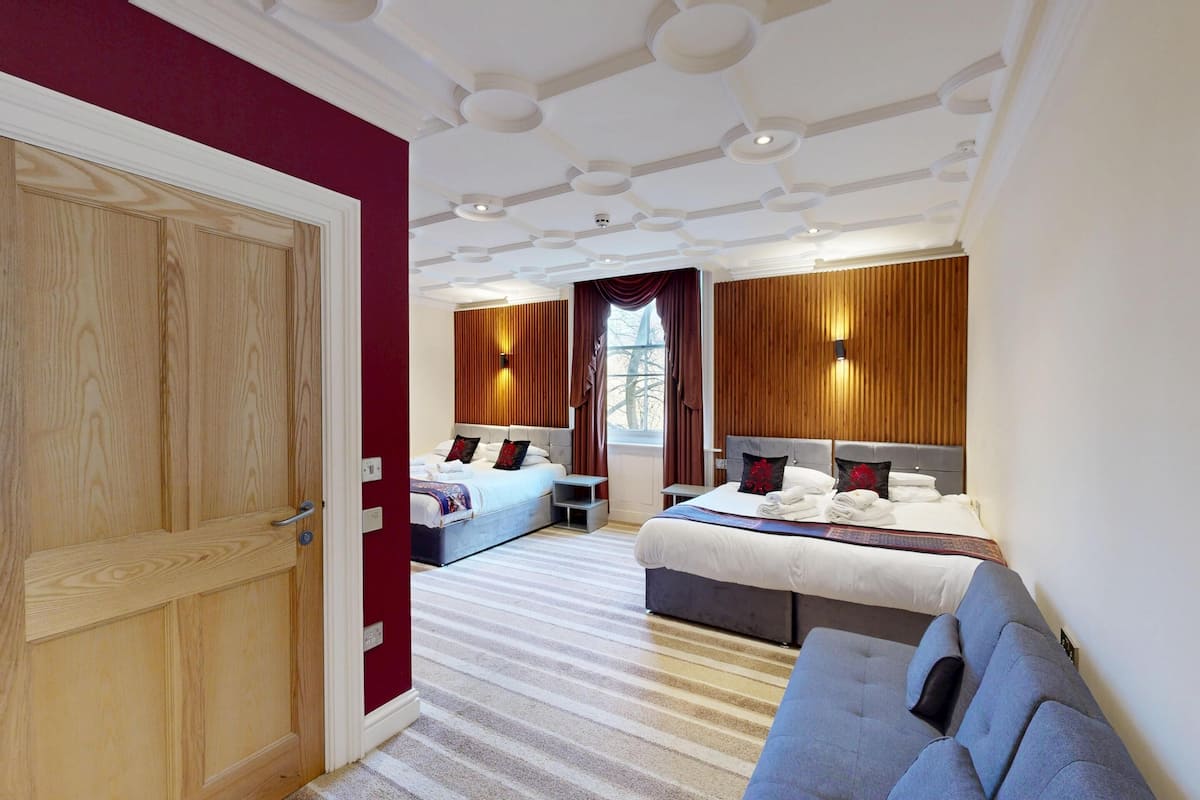A hotel room with two beds, a sofa, a wooden wardrobe, and a large window with curtains.