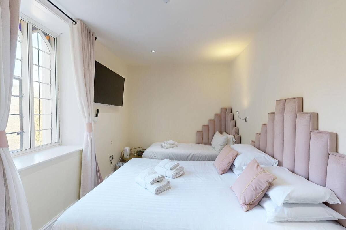 A hotel room with two beds, a large window with curtains, a flat-screen TV mounted on the wall, and a unique headboard design.