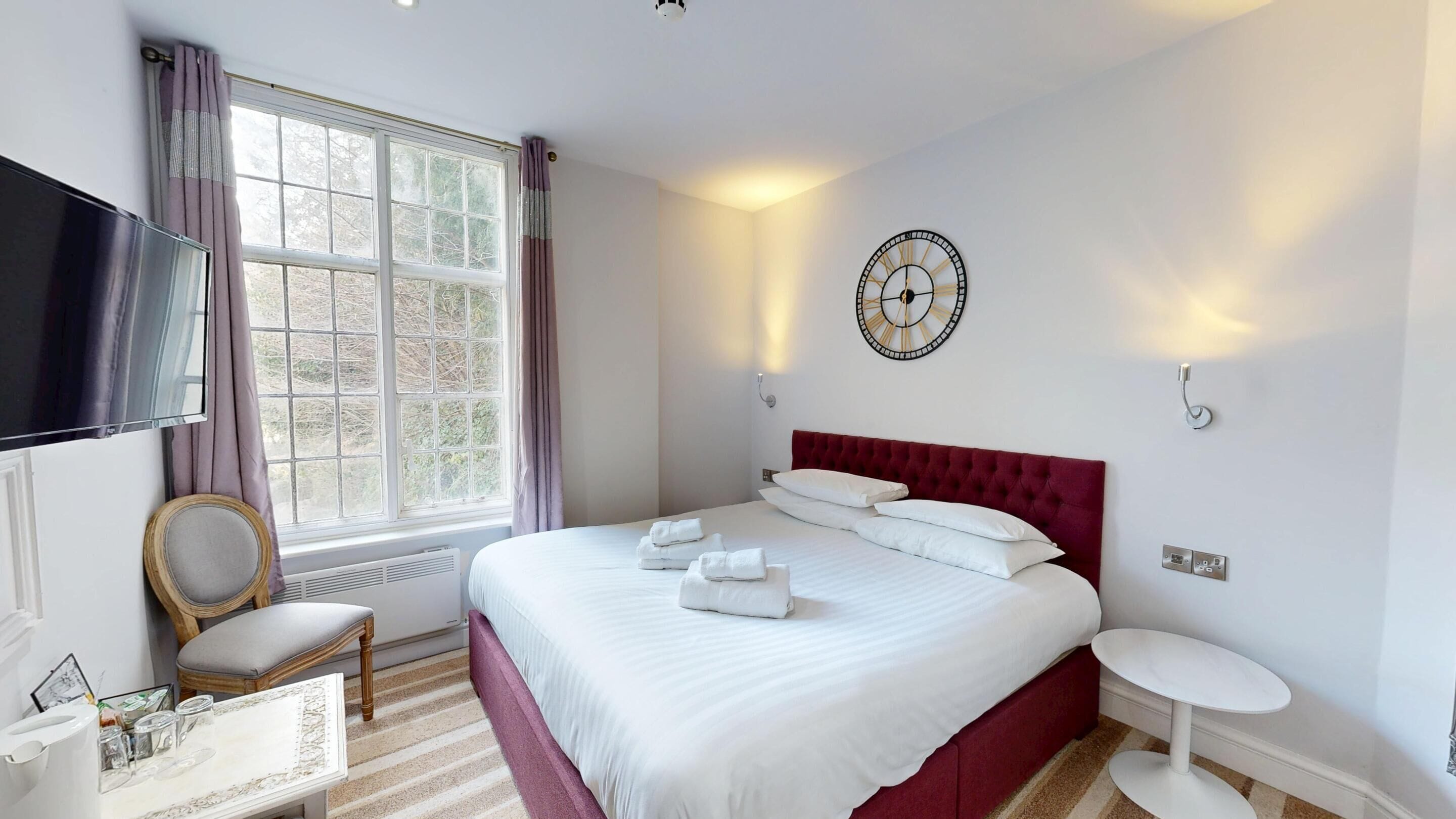 Una habitación de hotel con una cama grande, un televisor de pantalla plana, una ventana con cortinas, un reloj de pared y una pequeña mesa con una silla.