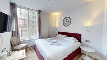 Hotelkamer met een groot bed, een flatscreen televisie, een raam met gordijnen, een wandklok en een klein tafeltje met een stoel.