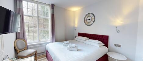 Una habitación de hotel con una cama grande, un televisor de pantalla plana, una ventana con cortinas, un reloj de pared y una pequeña mesa con una silla.