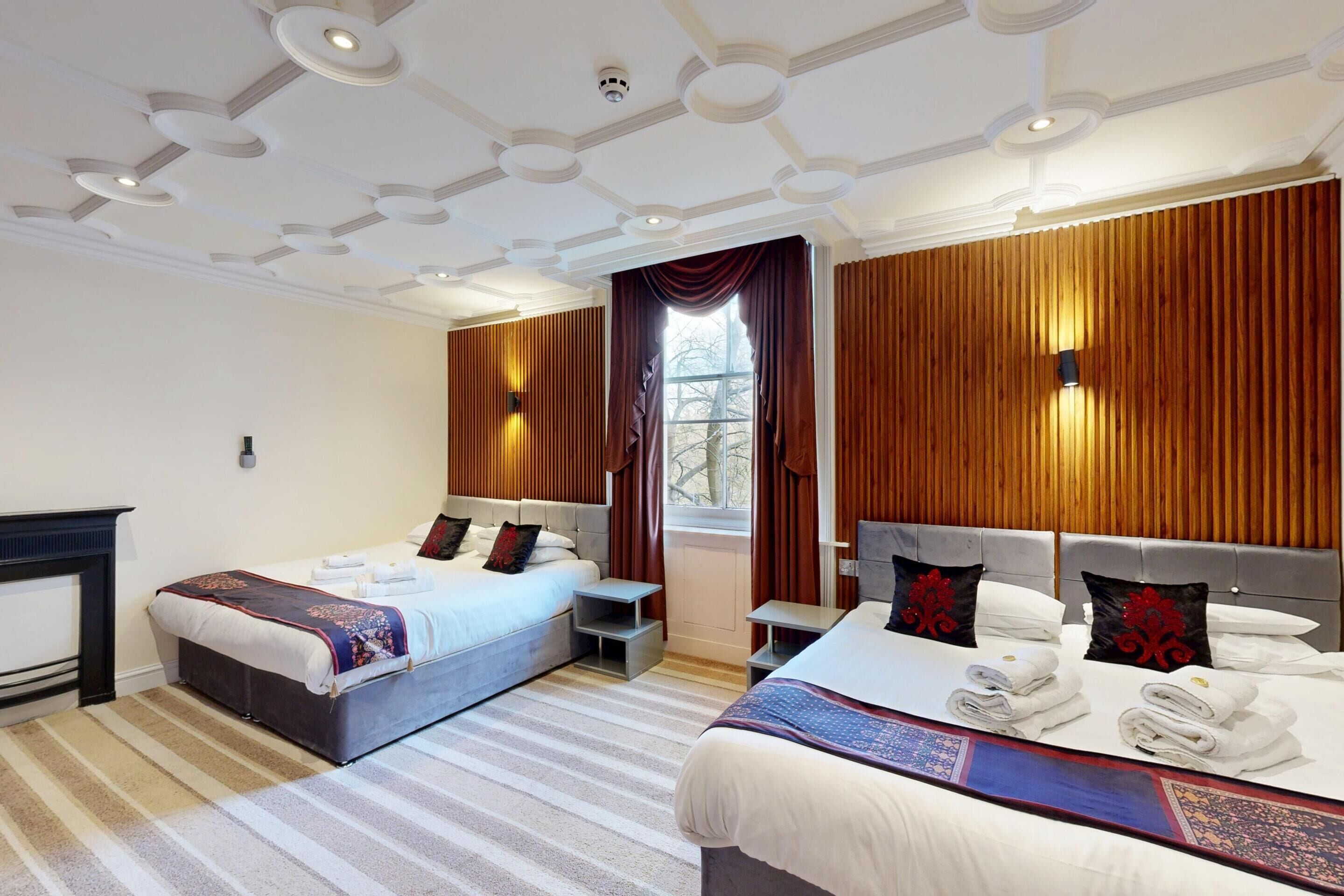 A hotel room with two beds, a fireplace, a large window with curtains, and a wooden wall paneling.
