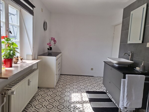 Un baño moderno con un lavamanos blanco, un espejo, un radiador, baldosas de suelo estampadas y una planta en maceta en el alféizar de la ventana.