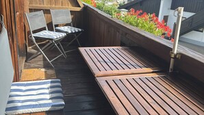 A wooden deck with two lounge chairs and a table, overlooking a scenic view of a forested hill and buildings.