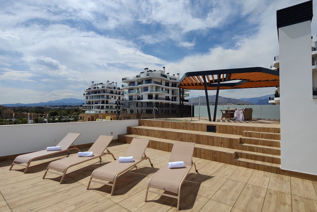 A rooftop terrace with sun loungers, a wooden deck, and a view of a multi-story building in the background.