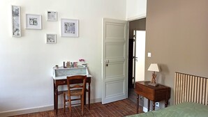 Una habitación con un escritorio de madera, una silla, una cama con cubrecama verde, una mesita de noche con lámpara y una puerta que da a otra habitación.