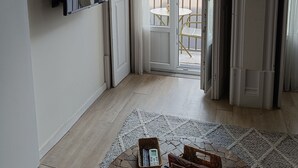 A room with a large window, a flat-screen TV mounted on the wall, a small table with a basket of books, and a wooden floor. - Porto Alma 388 - Silent City center (Porto)
