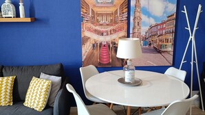 A room with a blue wall, a round white table with chairs, a sofa with patterned cushions, and two paintings depicting architectural scenes. - Porto Alma 388 - Silent City center (Porto)