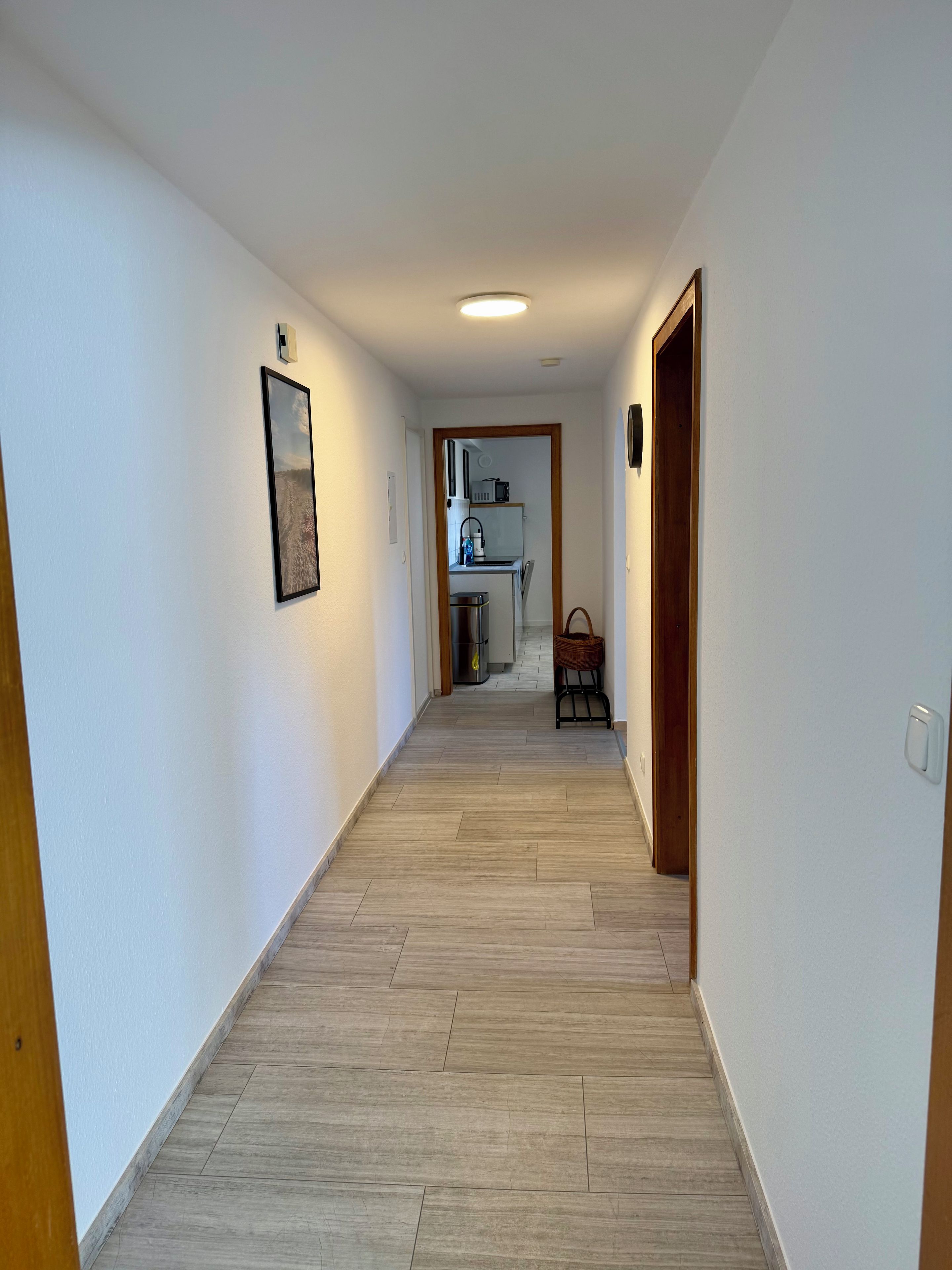 A hallway with a wooden floor, a framed picture on the left wall, and a kitchen area visible through an open doorway.