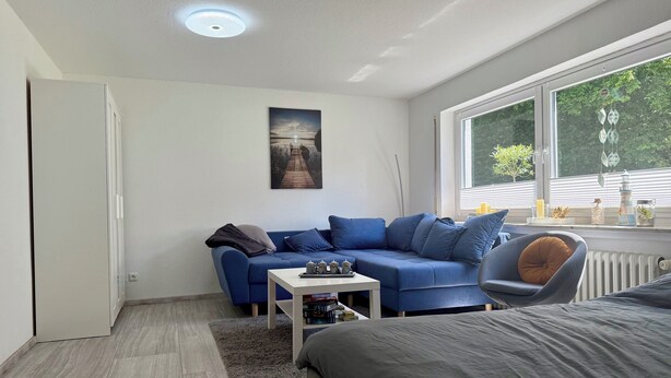 A modern, well-lit room with a blue sofa, a small table, a bed, and a large window offering a view of greenery outside. - Weserglück vacation apartment Grohnde (Emmerthal)