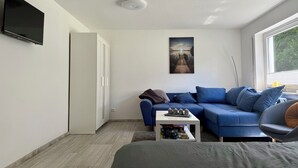 A modern, well-lit room with a blue sofa set, a flat-screen TV mounted on the wall, and a window providing natural light. - Weserglück vacation apartment Grohnde (Emmerthal)