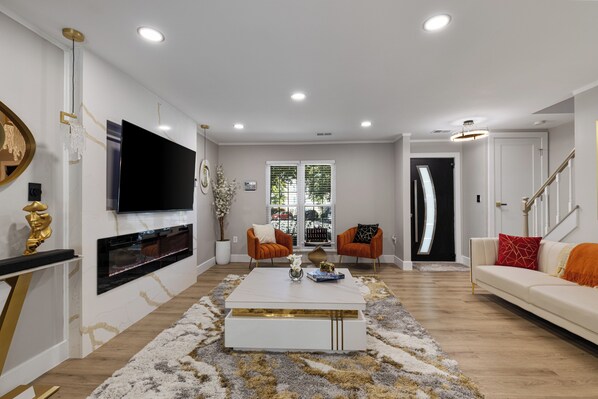 A modern living room with a flat-screen TV mounted on the wall, a white sofa, two armchairs, a coffee table, and a staircase leading to another level.