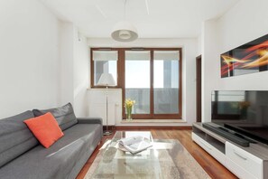 Uma sala de estar moderna com um sofá cinza, uma almofada vermelha, uma mesa de centro de vidro, um rack para TV e um quadro na parede.