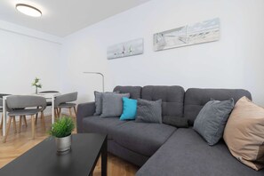 Sala de estar moderna com um sofá cinzento, uma mesinha preta e uma planta em vaso. Há uma área de jantar com uma mesa branca e cadeiras cinzas.