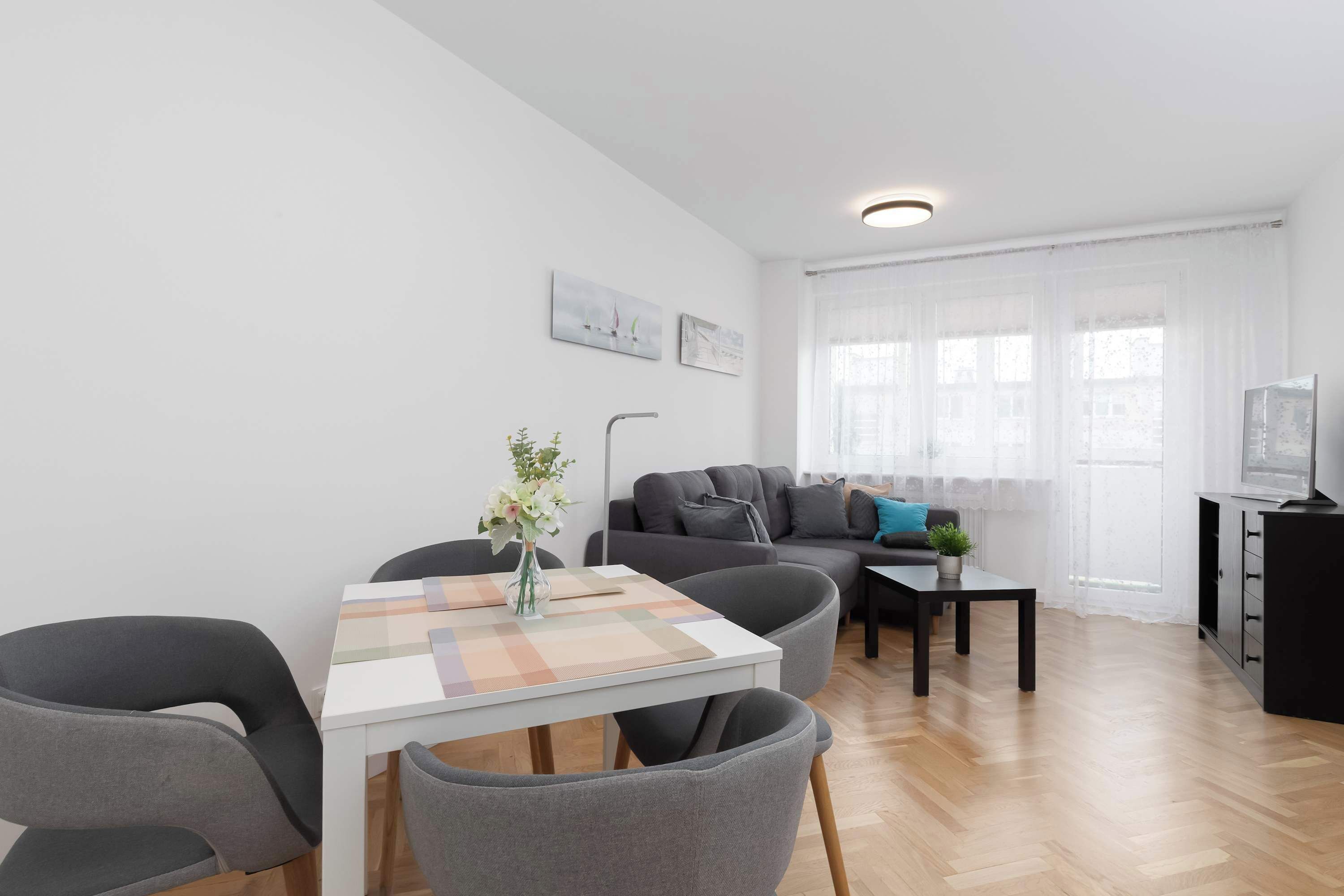 A modern living room with a sofa, a dining table with chairs, a small side table, and a TV on a cabinet.