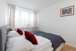 Quarto com cama, travesseiros brancos e vermelhos, cabeceira cinza, janela com cortinas leves e um quadro na parede.
