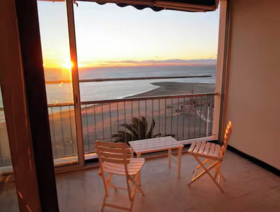 Un balcon donnant sur le coucher de soleil sur la mer, deux chaises en bois et une petite table.