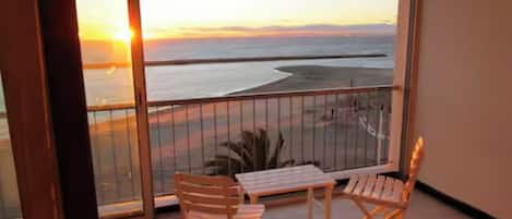 A balcony with a view of the sunset over the sea, two wooden chairs, and a small table.