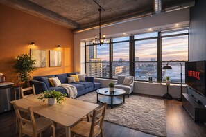 Una sala de estar moderna con sofá, mesa de centro y televisor, con amplias ventanas y vista a la ciudad.