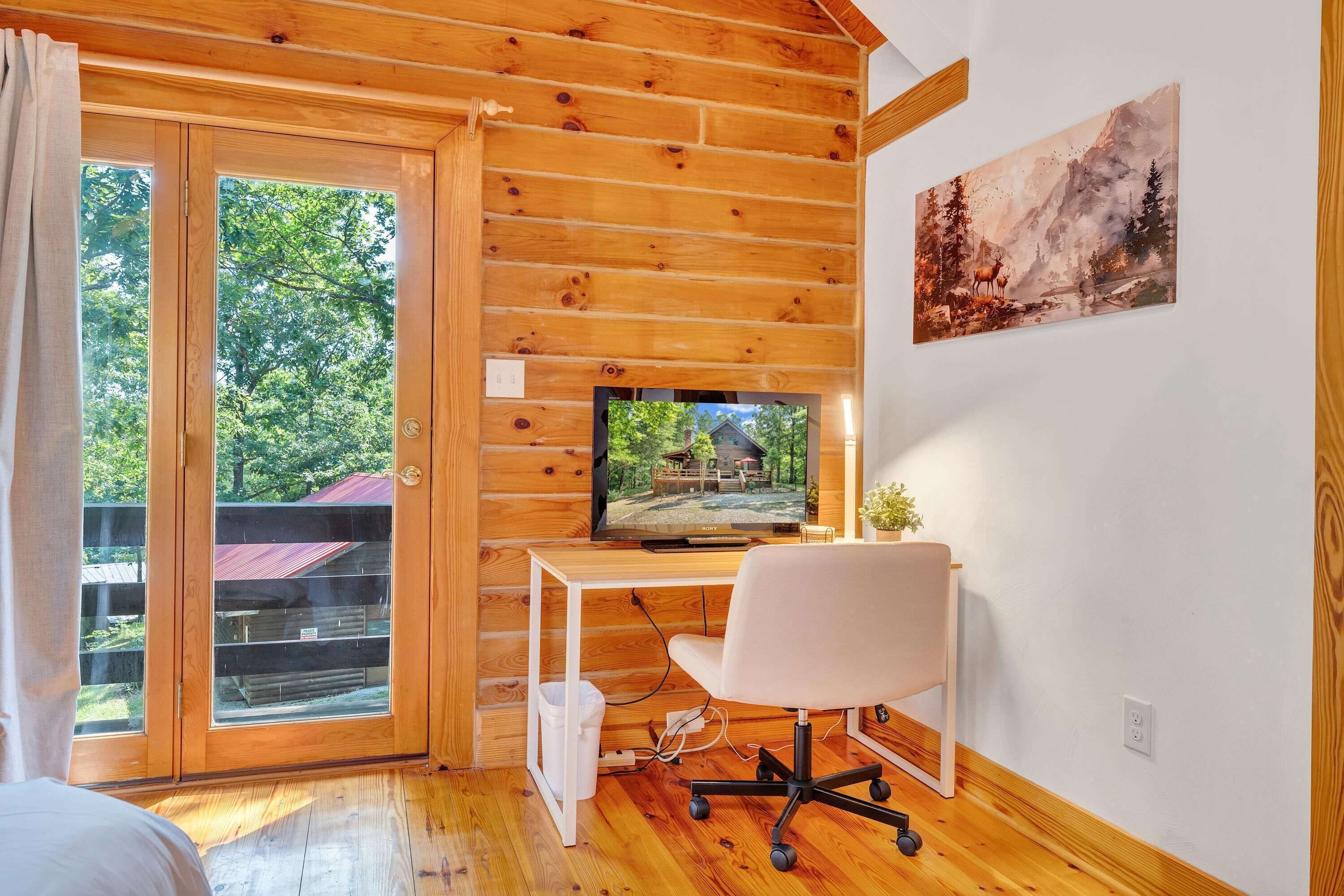 Ein Zimmer in einer Holzhütte mit Fernseher auf einem Schreibtisch, einem Sessel und einem Fenster mit Blick auf einen Wald.