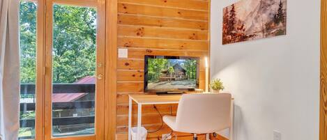 Ein Zimmer in einer Holzhütte mit Fernseher auf einem Schreibtisch, einem Sessel und einem Fenster mit Blick auf einen Wald.