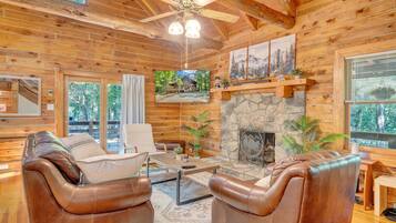 A log cabin interior with leather sofas, a stone fireplace, wooden walls, and a large TV screen.