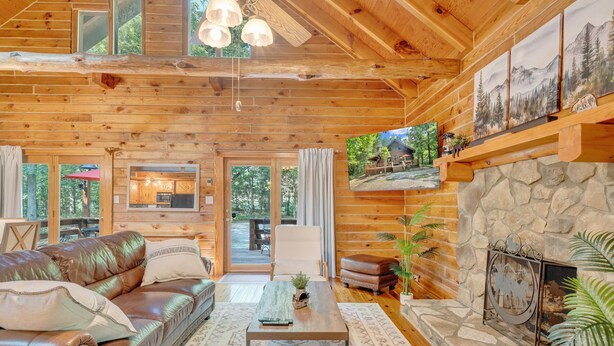 A cozy log cabin interior with a leather sofa, a stone fireplace, and a dining area visible through the glass doors. - Eagles Rest~ Grand, Luxurious & Peaceful— Near Hiking Trails & Virginia Tech! (New Castle)
