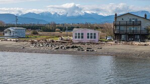 A coastal area with a pink building, a larger two-story house, and mountains in the background. - Oceanfront House w/ Deck, Firepit & Mountain Views (Sequim)