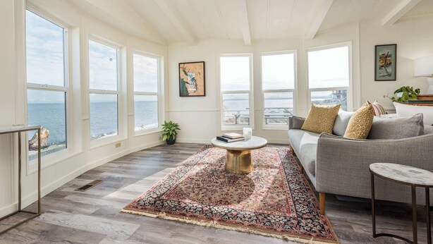 A spacious room with large windows offering ocean views, a grey sofa, a patterned rug, and a small round table. - Oceanfront House w/ Deck, Firepit & Mountain Views (Sequim)
