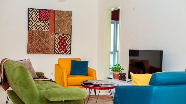 Een woonkamer met een groene fauteuil, een blauwe bank, een oranje fauteuil en een rode salontafel. Op de muur hangt een groot kunstwerk met een patroon en er is een televisie aan de muur bevestigd.