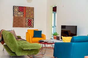 A living room with a green armchair, a blue sofa, an orange armchair, and a red coffee table. A large patterned artwork is on the wall, and a TV is mounted on the wall.