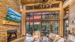 A spacious living room with a stone fireplace, large windows, and a ceiling fan. - Mountain Dream’n- modern, dog friendly, hot tub (Blue Ridge)