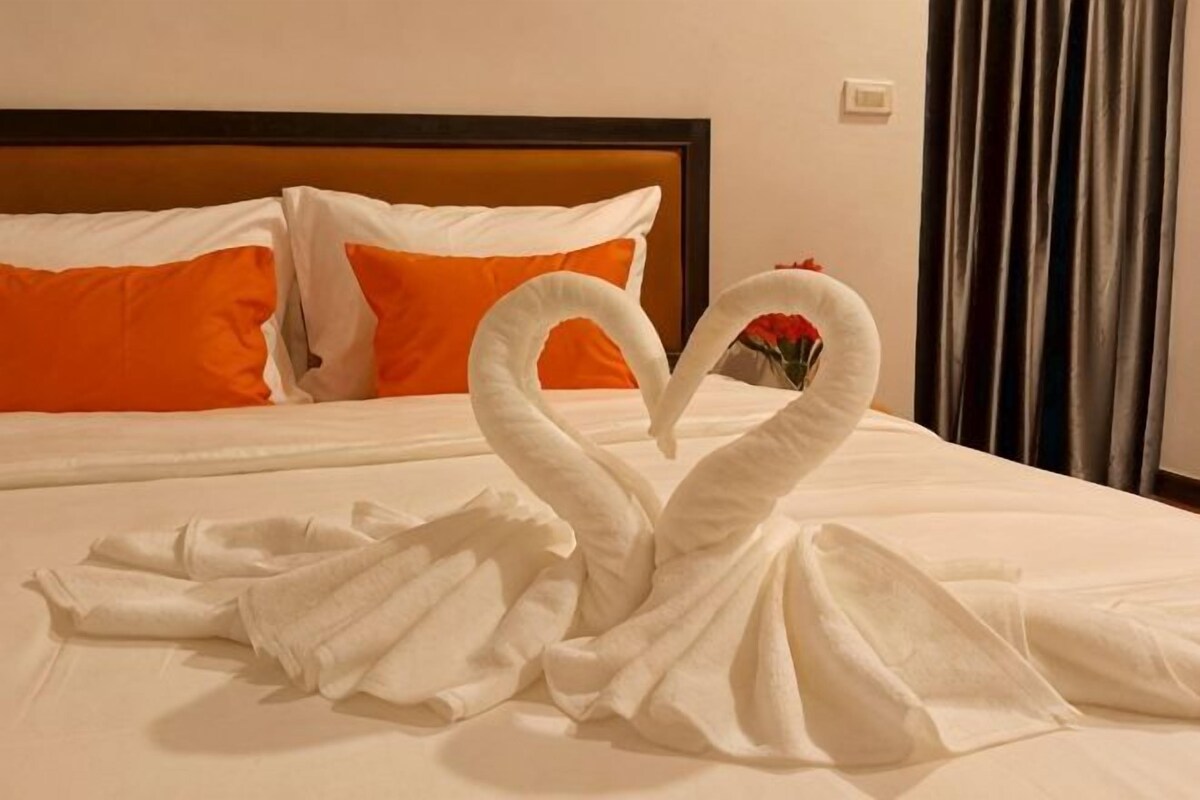 A bed with white and orange pillows, a heart-shaped towel arrangement, and a wooden headboard.