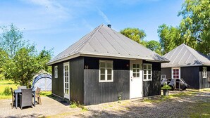A small, single-story building with a dark exterior and a sloped roof. It has a number '16' on the side, a door, and windows. There is a tent and outdoor seating area nearby. - 0 Person Holiday Park Home in Hesselager (Hesselager)