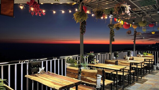 An outdoor seating area with wooden tables and chairs, hanging plants, and string lights against a sunset backdrop. - Lord Krishna Boutique Stay (Dharamshala)