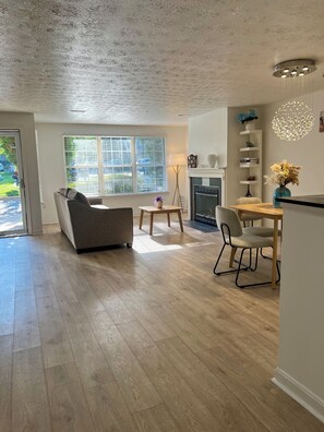 Ein modernes Wohnzimmer mit einer Couch, einem Esstisch mit Stühlen, einem Kamin und einem großen Fenster mit Jalousien.