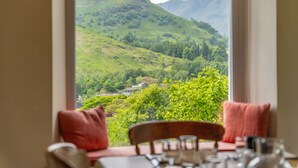 Un coin repas avec une table dressée pour deux personnes, et une fenêtre donnant sur une vallée verdoyante et des collines.