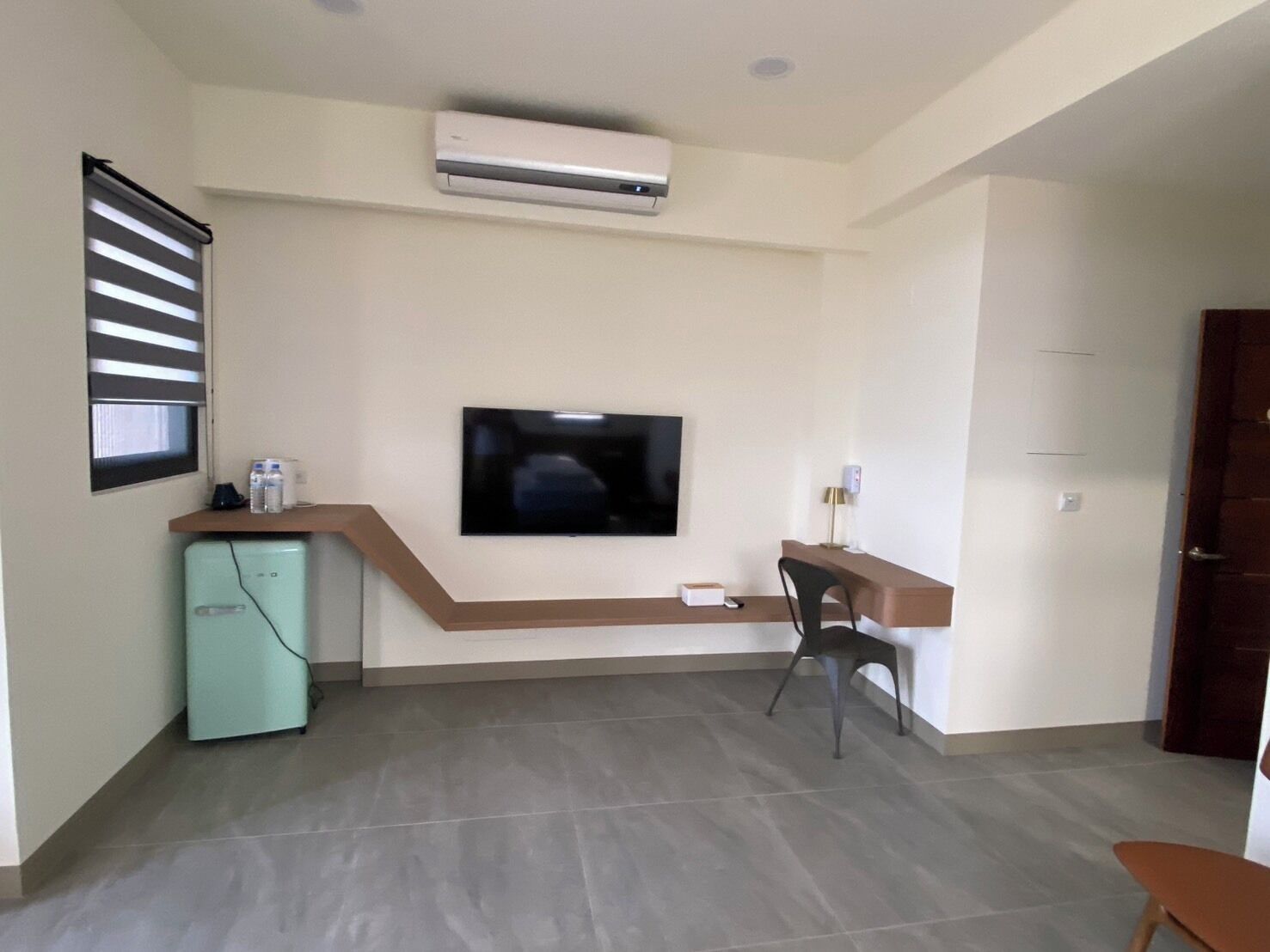 A compact living space with a flat-screen TV mounted on the wall, a small kitchenette with a fridge, and a seating area with a chair and table.