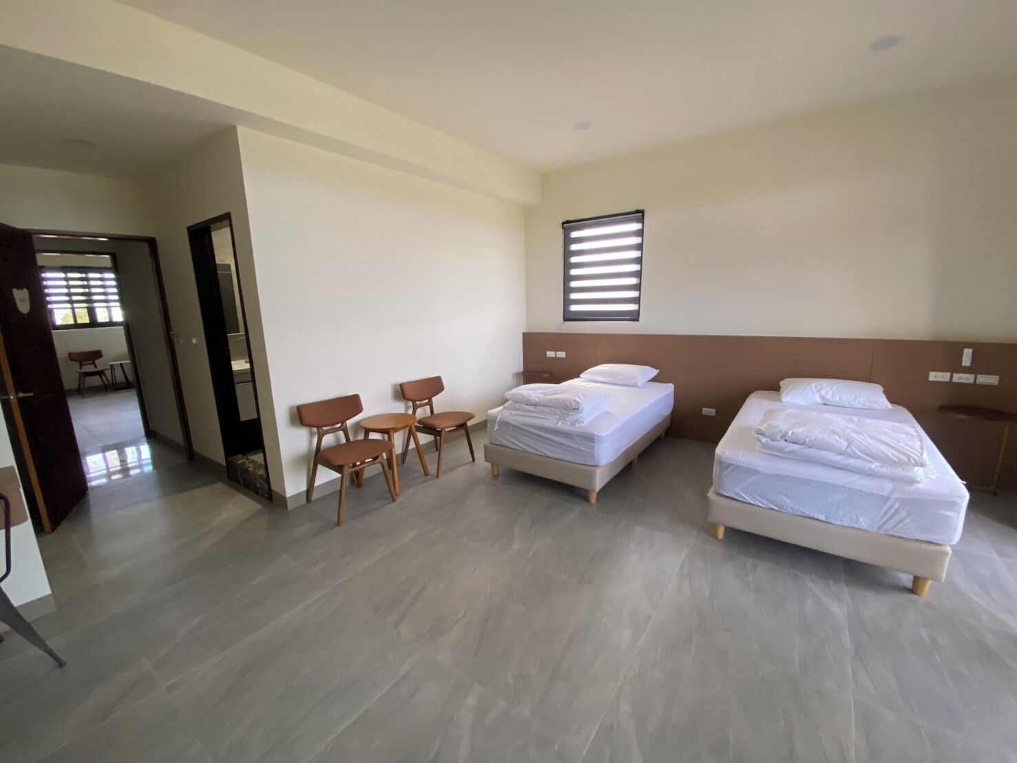 A hotel room with two beds, a small table with chairs, and a window with blinds.