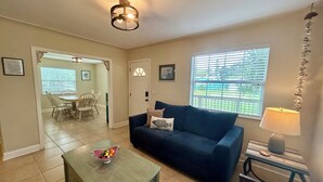 Un salon avec un canapé bleu foncé, une petite table basse verte, une lampe et une vue sur un jardin par une fenêtre.