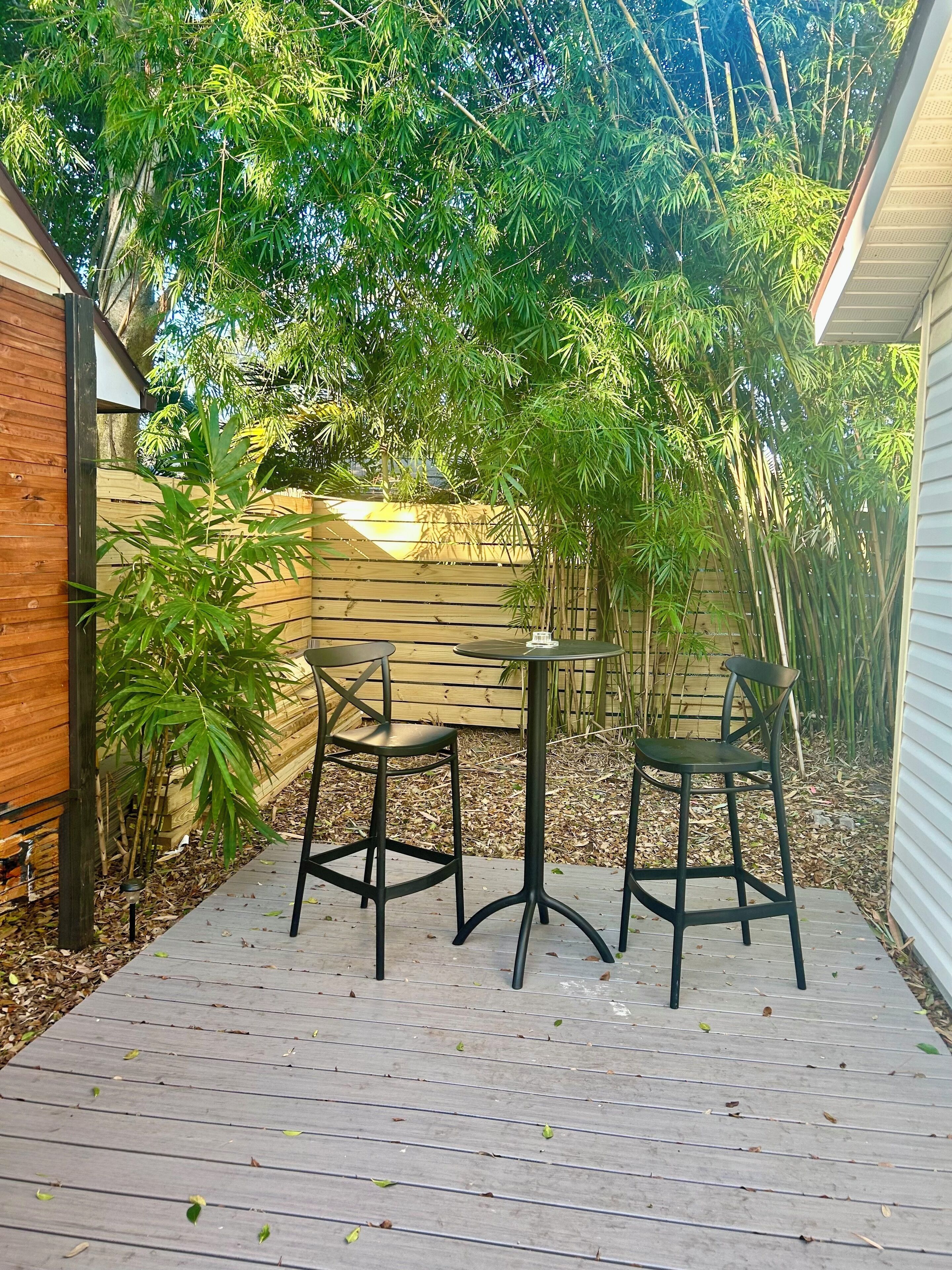 Une terrasse en bois avec deux chaises noires et une petite table ronde, entourée de plantes de bambou et d’une clôture en bois.